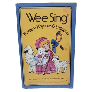 May include: A vintage "Wee Sing" book featuring a yellow cover with the title and subtitle "Nursery Rhymes & Lullabies" in black text. The cover art depicts a girl in a pink dress and bonnet with sheep. The book is in a blue frame.