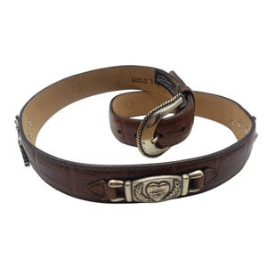 May include: Brown leather belt with a silver heart-shaped buckle and a silver buckle with the text "L 21207 MONTON." The belt has a crocodile-embossed pattern.