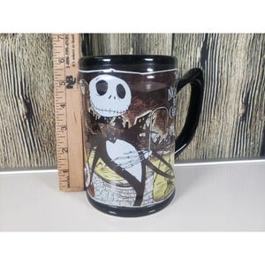 May include: Black ceramic mug featuring Jack Skellington from The Nightmare Before Christmas. The mug has a black handle and rim, with a detailed illustration of Jack and the text "Nightmare Before Christmas". The mug is approximately 5 inches tall.