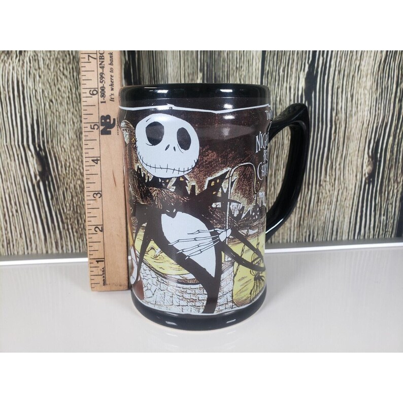 May include: Black ceramic mug featuring Jack Skellington from The Nightmare Before Christmas. The mug has a black handle and rim, with a detailed illustration of Jack and the text "Nightmare Before Christmas". The mug is approximately 5 inches tall.