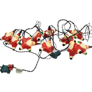 May include: A string of Christmas lights featuring red Santa Claus figures with white beards and hats. The lights are connected by black wires and have a green plug.