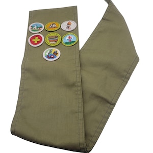 May include: Olive green sash with seven embroidered patches. The patches feature various colorful designs, including a church, animals, a campfire, a cross, a basket, a landscape, and a person with a boat. The sash is made of a durable fabric.