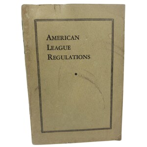 May include: A rectangular, tan-colored booklet with a thin black border frame. The front cover features the black text "American League Regulations" printed in a serif font, with a small black dot positioned below the text and a faint curved mark.