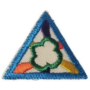 May include: A triangular embroidered patch featuring a white background with a central green four-leaf clover shape. The border is bright blue, and the design includes small orange, yellow, and dark purple accents within the triangular field.