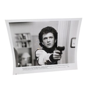 May include: Black and white promotional photo featuring a man holding a handgun, with a concerned expression. The image is a still from the movie "Lethal Weapon 2". The man is wearing a white shirt and a dark vest.