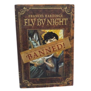 May include: A hardcover book titled "FLY BY NIGHT" by Frances Hardinge. The cover features a vintage design with a central illustration of a person holding a bird, with a "BANNED!" sticker across the image. The book has a brown and gold color scheme.