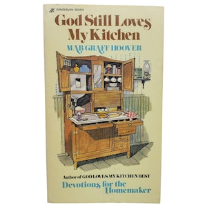 May include: A paperback book titled God Still Loves My Kitchen by Mab Graff Hoover. The cover features a brown ink illustration of a wooden kitchen hutch with open doors, shelves filled with jars and dishes, and a countertop with a rolling pin.