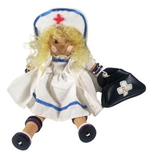May include: A wooden nurse doll with blonde curly hair, wearing a white uniform with blue trim and a matching cap with a red cross. The doll holds a black bag with a white cross and Rx symbol.
