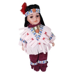 May include: A porcelain doll dressed in Native American attire. The doll has black hair, a headband with a flower, and a red feather. The doll wears a pink fringed top, burgundy pants, and a patterned scarf.