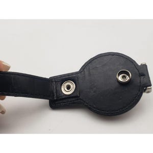 May include: Black leather accessory with a circular design and a strap. Features two silver snap closures and a metal clasp. The accessory is likely used for securing or fastening.
