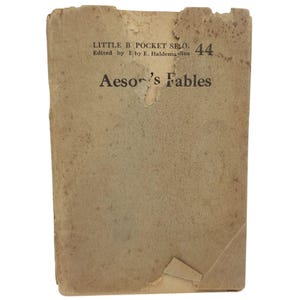 May include: A vintage, weathered tan paperback book titled Aesop's Fables, part of the Little Blue Book series, number 44. The cover shows significant wear, including frayed edges, a torn top section, and scattered brown age spots throughout.
