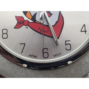 May include: Close-up of a silver-toned wristwatch with black numerals and hands. The watch face features a cartoon graphic with red, white, and orange accents. The text "TONY" is visible on the face, along with "JAPAN" and "MOVT".