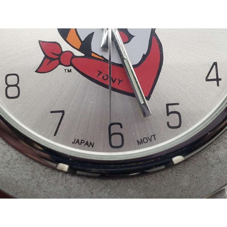 May include: Close-up of a silver-toned wristwatch with black numerals and hands. The watch face features a cartoon graphic with red, white, and orange accents. The text "TONY" is visible on the face, along with "JAPAN" and "MOVT".