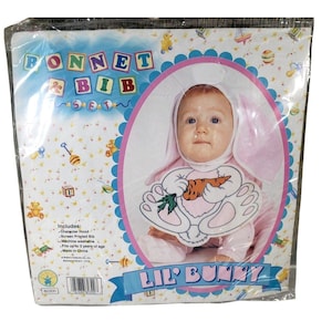 May include: A packaged Lil' Bunny costume set for babies and toddlers. The set includes a pink character hood with bunny ears and a screen-printed bib featuring a bunny holding a carrot. The packaging reads "Bonnet & Bib Set".