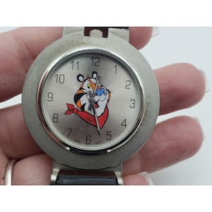 May include: A silver-toned wristwatch with a round face featuring the cartoon character Tony the Tiger. The watch has a black leather band and a silver bezel. The watch face displays the numbers 1 through 12.