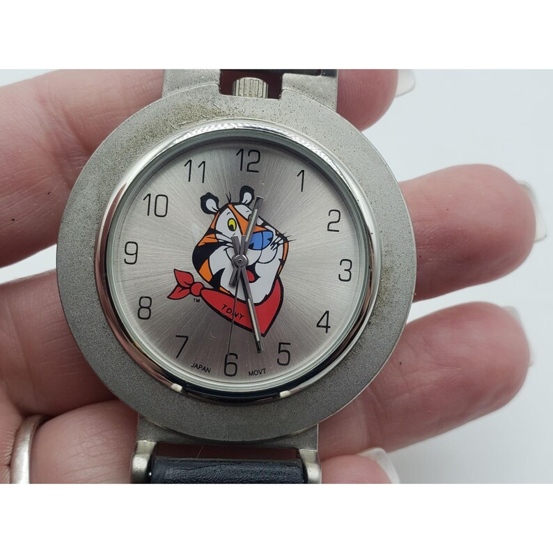 May include: A silver-toned wristwatch with a round face featuring the cartoon character Tony the Tiger. The watch has a black leather band and a silver bezel. The watch face displays the numbers 1 through 12.