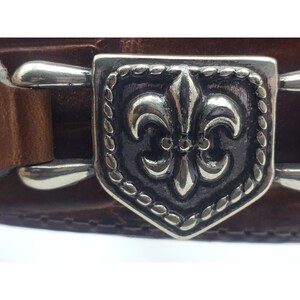 May include: A brown leather belt with a silver buckle featuring a fleur-de-lis design.