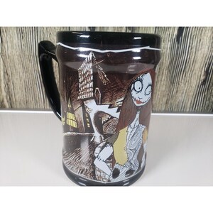 May include: Black ceramic mug featuring Sally from The Nightmare Before Christmas. The mug has a black handle and rim, with a detailed illustration of Sally and a spooky town scene. The mug is a standard size for hot beverages.