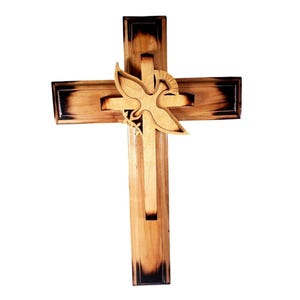 May include: Wooden cross with a dove and olive branch cutout. The cross is made of light-colored wood with darker, burnt edges. The dove is a lighter shade of wood, and the design is suitable for religious home decor.