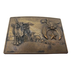May include: A brass belt buckle with a raised design of a camel caravan and a crescent moon and star. The buckle is a traditional Masonic symbol.