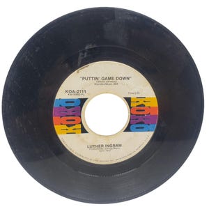 May include: A black vinyl record with a white and colorful label. The label features the text "PUTTIN' GAME DOWN" by Luther Ingram. The record is a 45 RPM single, produced by Johnny Baylor in April 1972.