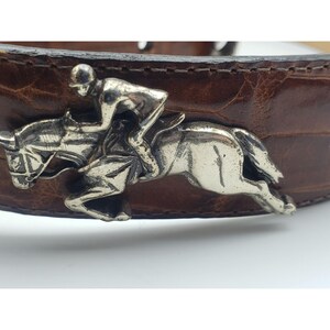 May include: A brown leather belt with a silver-toned buckle featuring a jockey riding a horse.