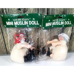 May include: Two "Just For Keeps" mini muslin dolls in plastic packaging. One doll is Santa Claus with a red hat and black boots. The other doll is a woman with a white bonnet and black shoes. The dolls are sitting down.