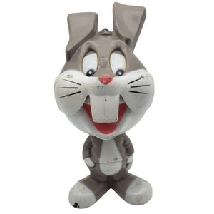 May include: A brown and white plastic rabbit figurine with large ears, a wide grin showing two prominent front teeth, and a red nose. The character has a cartoonish appearance with a rounded head, a small torso, and large feet, showing signs of wear.