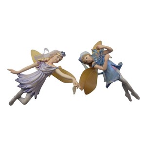 May include: Two fairies with wings, one in a lavender dress and the other in a blue dress, are flying and holding hands. They have blonde hair and are wearing crowns.