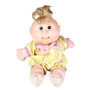 May include: A Cabbage Patch Kids doll with blonde hair tied in a top ponytail, featuring a round face, dark eyes, and a smiling expression. The doll wears a yellow romper with pink trim, a pink bow at the neckline, and a small pink logo on the chest.