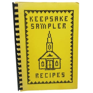 May include: A yellow spiral-bound cookbook titled "Keepsake Sampler Recipes." The cover features a black cross-stitch design of a church and border. The text is also in black cross-stitch.