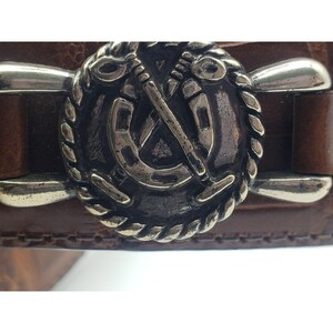 May include: A brown leather belt with a silver buckle featuring a horseshoe and crossed polo mallets.