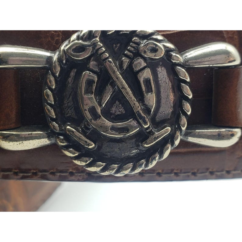 May include: A brown leather belt with a silver buckle featuring a horseshoe and crossed polo mallets.