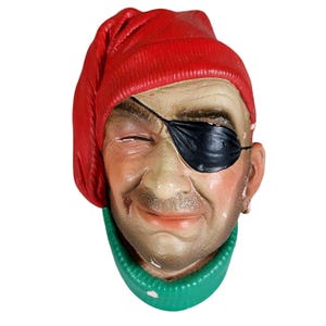 May include: A red and green ceramic figurine of a pirate with a black eye patch and a red hat. The pirate is smiling and has a mustache.