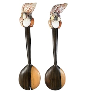 May include: A pair of salad servers featuring dark brown wooden handles topped with clusters of various small seashells, including a large spiral shell and a white fan-shaped shell. The spoon and fork bowls are made of two-toned dark and light wood.