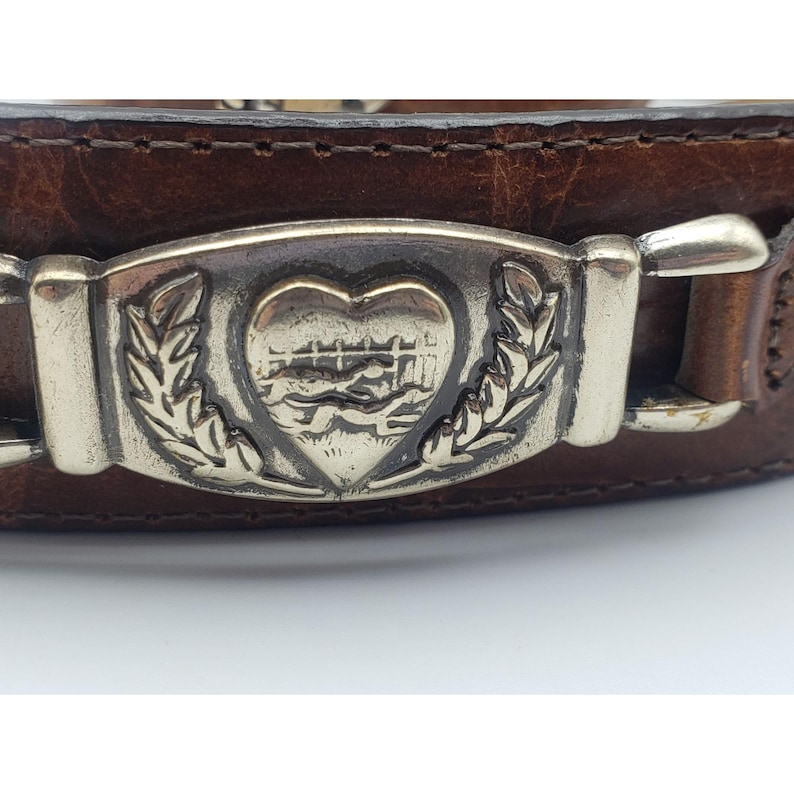 May include: A brown leather belt with a silver buckle featuring a heart with a stylized image of two figures inside. The heart is surrounded by two sprigs of leaves.