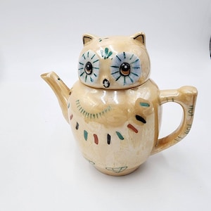 May include: A ceramic owl teapot with a light brown glaze and colorful painted details. The owl has large blue eyes with black pupils and a brown beak. The teapot has a handle and a spout.