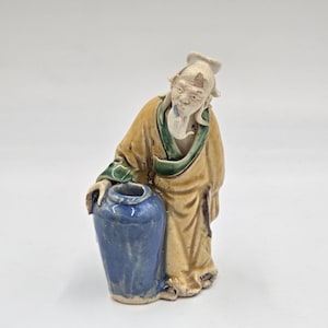 Op de afbeelding: Een keramieken beeldje van een oude man in een gele jurk met groene randen, leunend op een blauwe vaas. Het beeldje heeft een verweerde uitstraling, wat duidt op leeftijd en een vintage esthetiek. De man heeft een witte baard en een hoed.