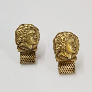 May include: A pair of gold-toned cufflinks featuring a circular coin-like design with a relief profile of a figure with curly hair. Each piece is attached to a rectangular, textured mesh-style metal base, creating a cohesive set of accessories.