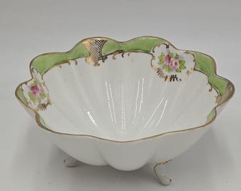 May include: A white porcelain bowl with a scalloped edge and three feet. The bowl is decorated with green and gold accents and floral designs.