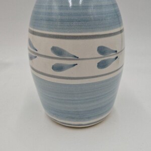 May include: A small, light blue ceramic vase with a white base and a gray stripe. The vase has a simple design with blue leaf-like shapes on the white stripe.