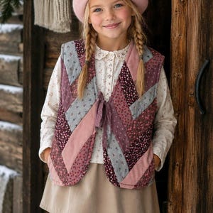 May include: A quilted vest featuring a chevron pattern in shades of dusty pink, mauve, and grey with small floral prints. The vest has a tie-front closure and is worn over a long-sleeved cream-colored blouse with a lace collar and a plain skirt.