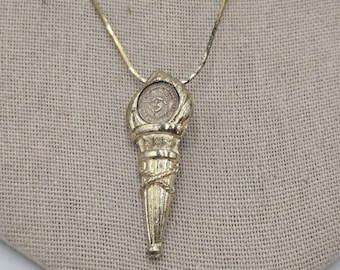 Könnte beinhalten: Goldkette mit einem goldenen Fackel-Anhänger, der eine runde Münze mit einem Design im Inneren trägt.