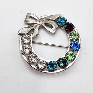 May include: A silver tone brooch in the shape of a wreath with a bow at the top. The wreath is decorated with colorful rhinestones in shades of blue, green, and red.