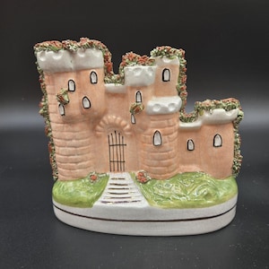 Puede incluir: Una figurita de cerámica de un castillo con un exterior de ladrillo rosa, hiedra verde y detalles blancos. El castillo tiene una pequeña puerta y un conjunto de escalones que conducen a ella.