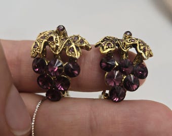 Könnte beinhalten: Ein Paar goldfarbene Ohrclips mit einem Traubenmotiv, besetzt mit violetten Strasssteinen.