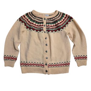 Könnte beinhalten: Cremefarbene Strickjacke mit rundem Ausschnitt und langen Ärmeln. Der Pullover hat ein detailliertes Muster in Rot und Grün um den Halsausschnitt, die Manschetten und den Saum. Er hat eine Knopfleiste mit silberfarbenen Knöpfen.