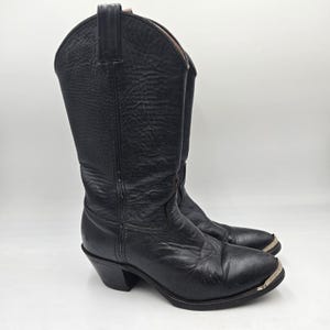 May include: A pair of black leather cowboy boots. The boots have a pointed toe, a stacked heel, and a metal accent on the toe. The shaft of the boots is tall, and the leather has a textured appearance. The boots are set against a white background.