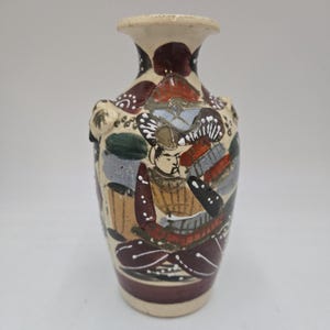 Könnte beinhalten: Eine kleine, cremefarbene Keramikvase mit rundem Körper und schmalem Hals. Die Vase ist mit einer farbenfrohen handgemalten Szene eines Samurai-Kriegers in Rüstung verziert, umgeben von Blumen und Blättern.