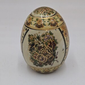 May include: An egg-shaped decorative container with a cream-colored base, adorned with floral and gold accents. The top and bottom feature intricate gold patterns, while the center showcases a detailed floral design with various colors.